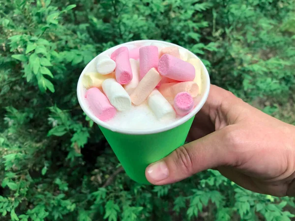 Coffee in a Green Paper Cup with whipped Cream and marshmallows on the green leaves background. Hand hold cup of milk drink with pink zephyr.