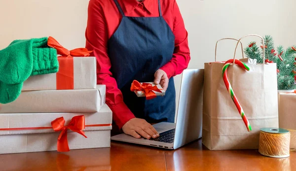 Deliver\'s Hands using Laptop near Packing bag, Gift\'s boxes and Christmas Tree. Online Delivery concept.
