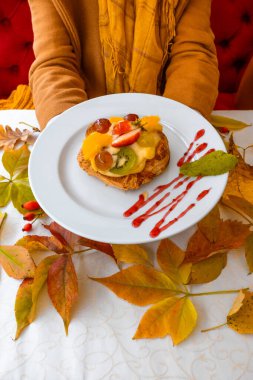 Sonbahar yaprakları üzerinde meyveli kek olan beyaz tabağı tutan kadın eller..