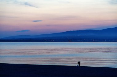 Sahilde yalnız bir figür günbatımında. Dramatik günbatımı, bulutlar, deniz, yalnızlık, dalgınlık, yansımalar yansımalar