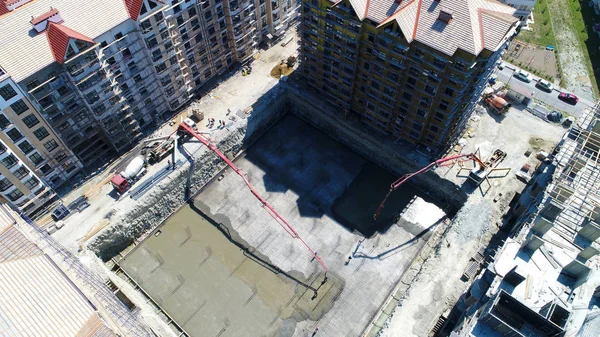 the casting of the concrete Foundation of a house. Bird's-eye view. Two concrete pumps feed the solution into an earthen pit, where workers level the concrete. Concrete is transported in a concrete mixer. Around are already built multi-storey houses