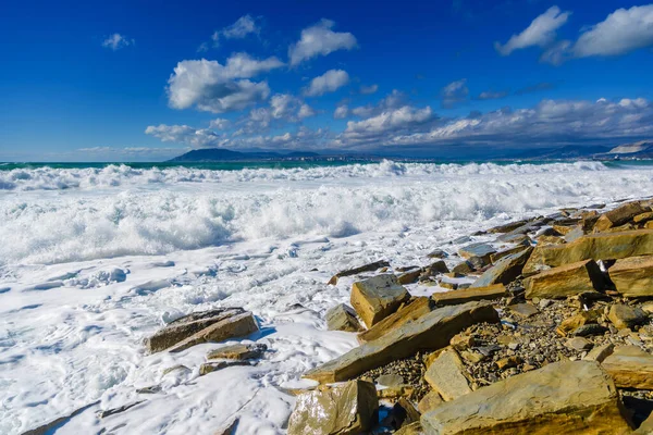 Storm in Tsemesskaya Bay. Turkoois golven met prachtig sneeuwwit schuim vallen op Cape Doob. Er liggen grote geelachtige rotsblokken aan de kust. Aan de horizon Novorossiysk en de bergen — Stockfoto