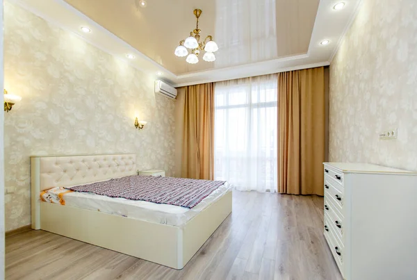 Room in bright colors with a new renovation. A large white bed and a white chest of drawers opposite the bed.Floor-length window with beige curtains