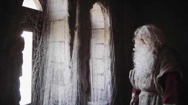 Старик с длинной белой бородой в красном халате смотрит в окно за пределами замка — стоковое видео