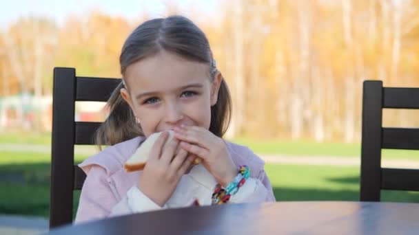 Chica divertida cinco años comiendo un sándwich en el parque en un café — Vídeos de Stock