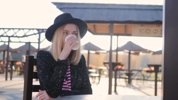 Una mujer con estilo en un sombrero está tomando café en un café en el parque. Ella sonríe, trae una taza con una bebida a su boca y la bebe . — Vídeo de stock