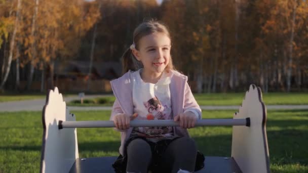 Маленька мила дівчинка гойдається і грає на ігровому майданчику в парку — стокове відео