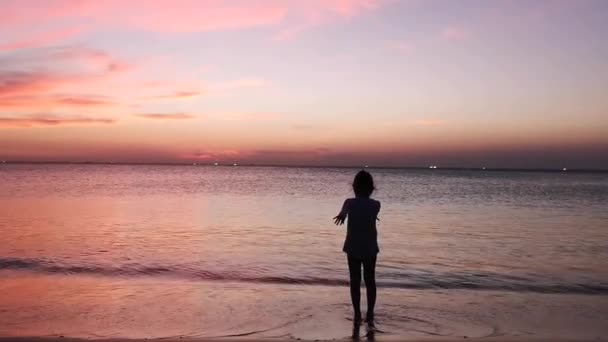 Силуэт маленькой девочки, машущей руками на закате у моря на пляже — стоковое видео