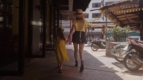 La petite fille dans la robe jaune va à la poignée avec sa mère en bas de la rue. Aimer le mode de vie de famille un jour ensoleillé. Vue de l'arrière — Video