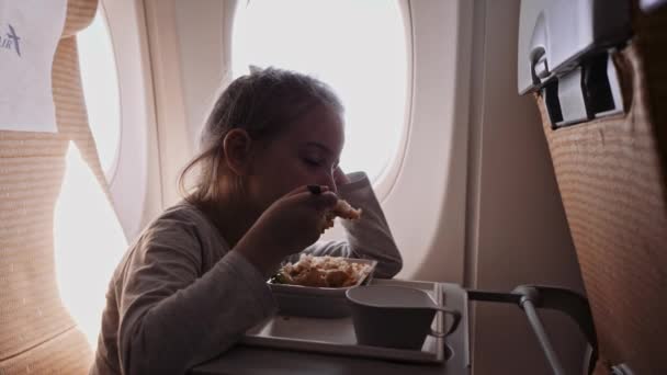 El niño está volando en un avión para descansar. Una chica sentada cerca de la ventana y comiendo — Vídeos de Stock