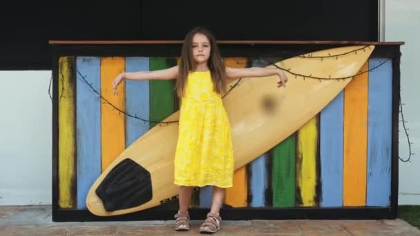 Μικρό όμορφο κορίτσι σε κίτρινο φόρεμα στέκεται κοντά στο Board surf — Αρχείο Βίντεο