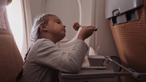 El niño está volando en un avión para descansar. Una chica sentada cerca de la ventana y comiendo — Vídeos de Stock