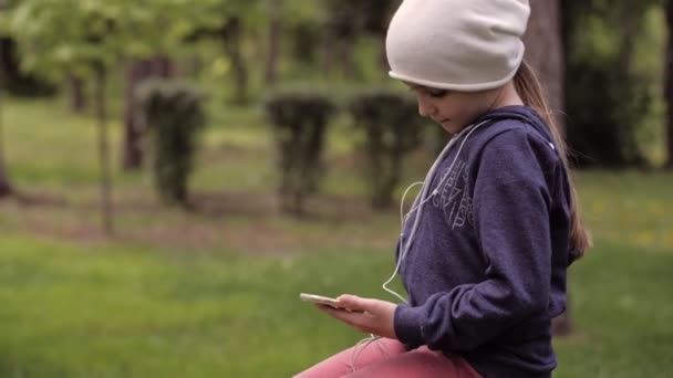 4K. Ένα χαριτωμένο κοριτσάκι που απολαμβάνει τη μουσική με έξυπνο τηλέφωνο στο πάρκο. Μόνος — Αρχείο Βίντεο