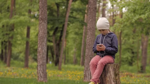 4K. Una niña linda disfrutando de la música con el teléfono inteligente en el parque. Solo. — Vídeo de stock