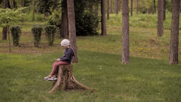 4K. Ένα χαριτωμένο κοριτσάκι που απολαμβάνει τη μουσική με έξυπνο τηλέφωνο στο πάρκο. Μόνος — Αρχείο Βίντεο