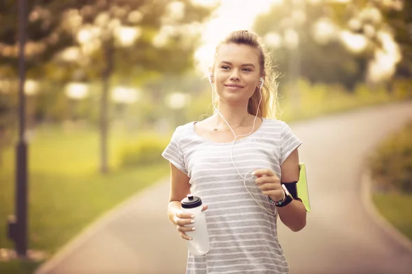 Young woman running in park and listening to music with headphones - Beautiful blonde and fit girl jogging alone outdoor in early autumn season - Front view