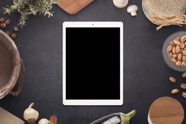 Tablet with blank screen on old country kitchen table surrounded with meal ingredients and kitchen items.