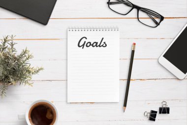 Goals list notepad mockup on office desk surrounded with smart phone, glasses, cup of coffee, pen, plant. Flat lay.