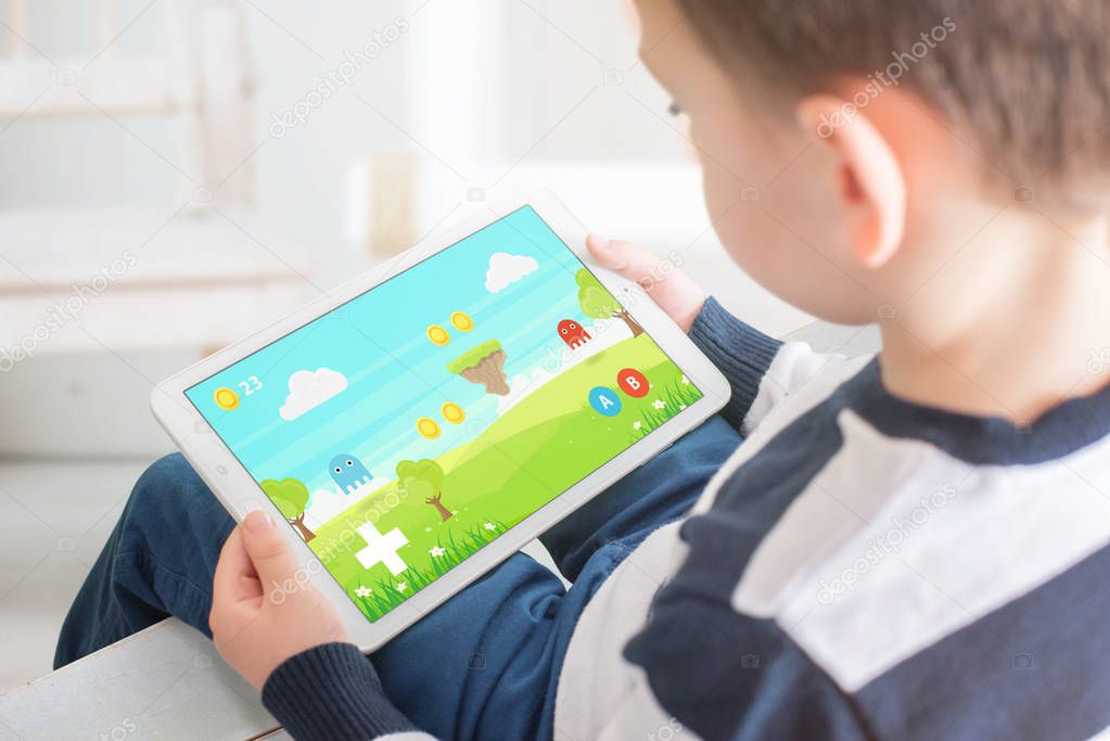 Boy playing game on white tablet. Home interior in bacgkround. Kid games on mobile devices concept.