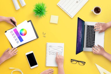 Web tasarım projesinde ekip çalışması. Üst görünüm, dizüstü bilgisayar, akıllı telefon, planlar, kağıtlar ve kahve ile düz yatıyordu sahne. Sarı masa.