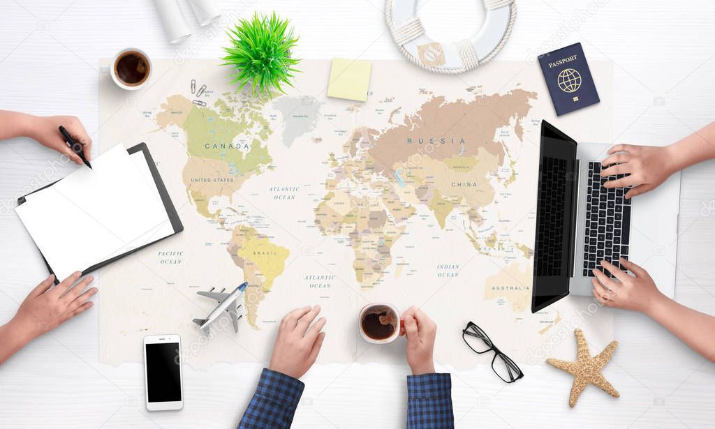 People are planning a holiday trip. Top view of the working tourist agency desk with a large world map in the background. Laptop, passport, papers, phone, glasses, and souvenirs on the desk.