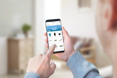 Adam akıllı telefonla evden alışveriş. Çevrimiçi mağaza uygulaması veya web sitesi kavramı. Arka planda oturma odası.