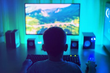 Çocuk bilgisayarda çalışıyor. Işıklı karanlık bir sahne. Arkadan bak, üçüncü şahıs. Genç bir hacker kavramı.