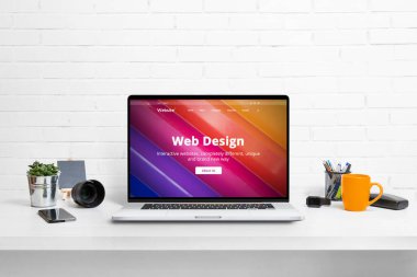 Dizüstü bilgisayarda web tasarımı konsepti web sitesi. Ofis masasında malzeme, bitki, kamera lensi, telefon, wathc ve kahve kupası var.