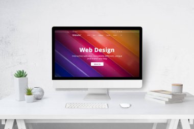 Çalışma masasında bilgisayar olan web tasarım stüdyosu konsepti ve sergilenen şirket web sitesi. Klavyesi, musluğu, bitkileri ve kitapları olan beyaz ahşap masa.