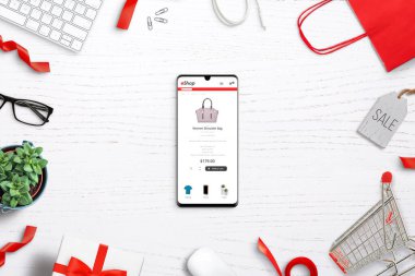 Akıllı telefon konseptiyle alışveriş yapmak. Telefon ekranında kadın çantası olan bir internet sitesi. Üst görünüm kompozisyonu beyaz masada alışveriş malzemeleriyle