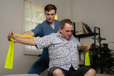 Fizyoterapist, klinikte direniş bandı, rehabilitasyon ve egzersiz kullanan yaşlı bir adama yardım ediyor..