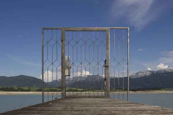 Looking through a locked lattice door onto a wooden boat dock across the dry Forgensee in the direction of the Alps
