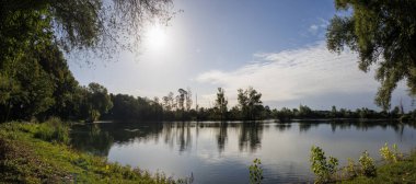 17 Eylül 2025 'te Almanya, Mering' de güneş ve ağaçlarla kaplı manzaralı göl manzarası