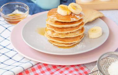 Ev yapımı muzlu krep, üzerine toz ve bal serpiştirilmiş. Mutfak masasındaki gözlemeler, arka plandaki yumurta ve fincanların yanında kahve. Ballı krep ve taze muzlu.