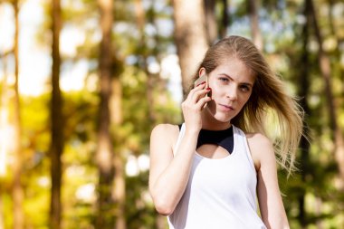 Güzel sarışın model spor giysileri cep telefonundan konuşuyor. Gün batımında bulanık orman arka plan