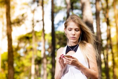 Güzel sarışın model cep telefonu müzik dinlerken spor giysiler içinde. Gün batımında bulanık orman arka plan