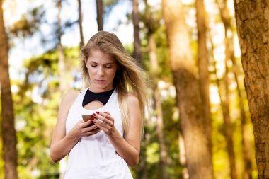 Güzel sarışın model cep telefonu kullanarak spor giysileri ile. Gün batımında bulanık orman arka plan