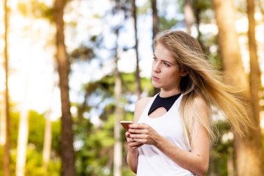 Güzel sarışın model cep telefonu kullanarak spor giysileri ile. Gün batımında bulanık orman arka plan