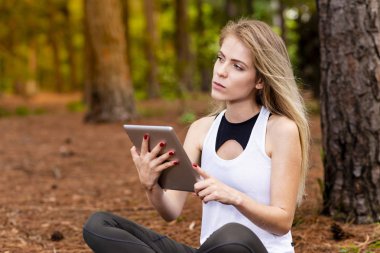 Kuru üzerinde oturan sarışın model okuma onun tablet üzerinde sopa. Gün batımında bulanık arka plan