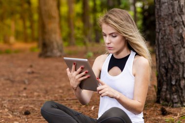 Kuru üzerinde oturan sarışın model okuma onun tablet üzerinde sopa. Gün batımında bulanık arka plan