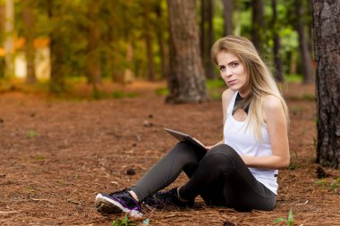 Kuru üzerinde oturan sarışın model okuma onun tablet üzerinde sopa. Gün batımında bulanık arka plan