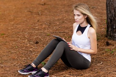 Kuru üzerinde oturan sarışın model okuma onun tablet üzerinde sopa. Gün batımında bulanık arka plan