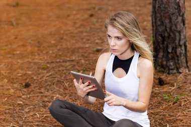 Kuru üzerinde oturan sarışın model okuma onun tablet üzerinde sopa. Gün batımında bulanık arka plan