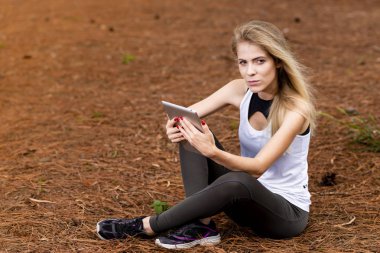 Kuru üzerinde oturan sarışın model okuma onun tablet üzerinde sopa. Gün batımında bulanık arka plan