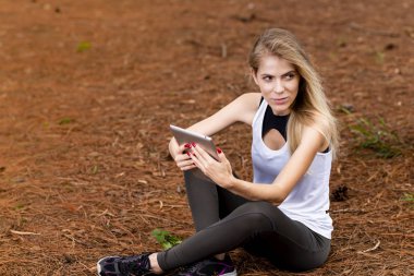 Kuru üzerinde oturan sarışın model okuma onun tablet üzerinde sopa. Gün batımında bulanık arka plan