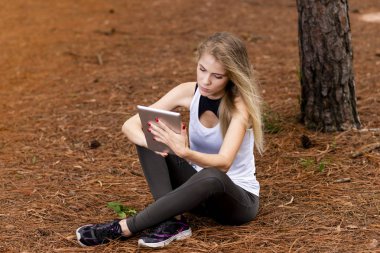 Kuru üzerinde oturan sarışın model okuma onun tablet üzerinde sopa. Gün batımında bulanık arka plan