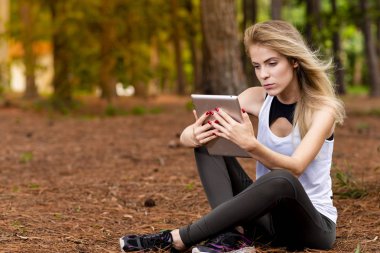 Kuru üzerinde oturan sarışın model okuma onun tablet üzerinde sopa. Gün batımında bulanık arka plan