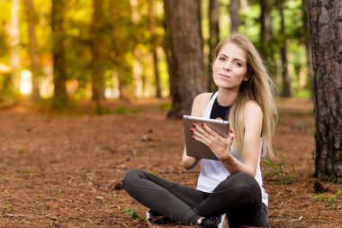 Kuru üzerinde oturan sarışın model okuma onun tablet üzerinde sopa. Gün batımında bulanık arka plan