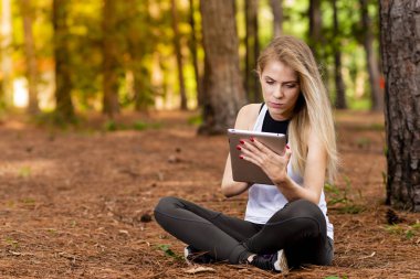 Kuru üzerinde oturan sarışın model okuma onun tablet üzerinde sopa. Gün batımında bulanık arka plan