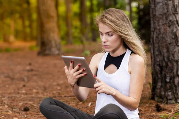 Kuru üzerinde oturan sarışın model okuma onun tablet üzerinde sopa. Gün batımında bulanık arka plan